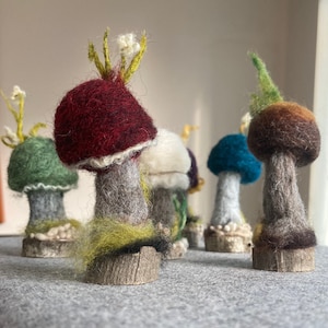 Puede incluir: Una colección de cinco figuritas de setas de fieltro con sombreros y tallos de diferentes colores. Las setas están dispuestas sobre una superficie gris y tienen un aspecto artesanal y caprichoso.