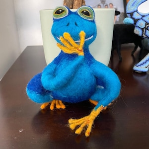 Puede incluir: Una figura de rana azul hecha con fieltro de aguja con ojos grandes y expresivos que presentan detalles en verde y negro. La rana tiene dedos y pies de color naranja brillante, con una mano levantada hacia la boca. La textura es suave y vellosa.