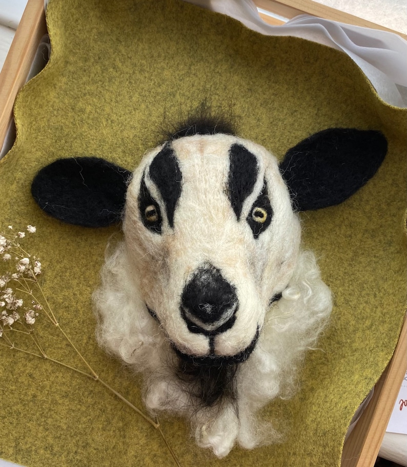 Puede incluir: Una cabeza de animal de fieltro blanco y negro con nariz negra y ojos amarillos. La cabeza del animal est&aacute; montada sobre un fondo de fieltro verde.