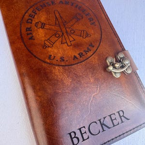 May include: A brown leather journal with the text "BECKER" engraved at the bottom. The journal features a circular emblem with the words "AIR DEFENSE ARTILLERY" and "U.S. ARMY" surrounding a crossed cannon and missile design. A metal clasp secures the journal.