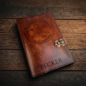 May include: A brown leather journal with a decorative clasp. The cover features a circular emblem with the text "AIR DEFENSE ARTILLERY U.S. ARMY" and the name "BECKER" engraved below the emblem.