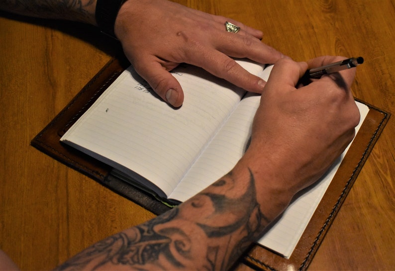 May include: A person with tattoos on their arms is writing in a brown leather bound journal with lined pages.