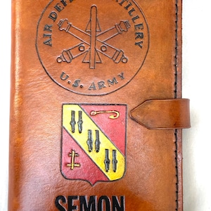 May include: A brown leather wallet with embossed text and imagery. The top features a circular emblem with "AIR DEFENSE ARTILLERY" and "U.S. ARMY". Below is a crest and the name "SEMON". The wallet has a leather strap closure.
