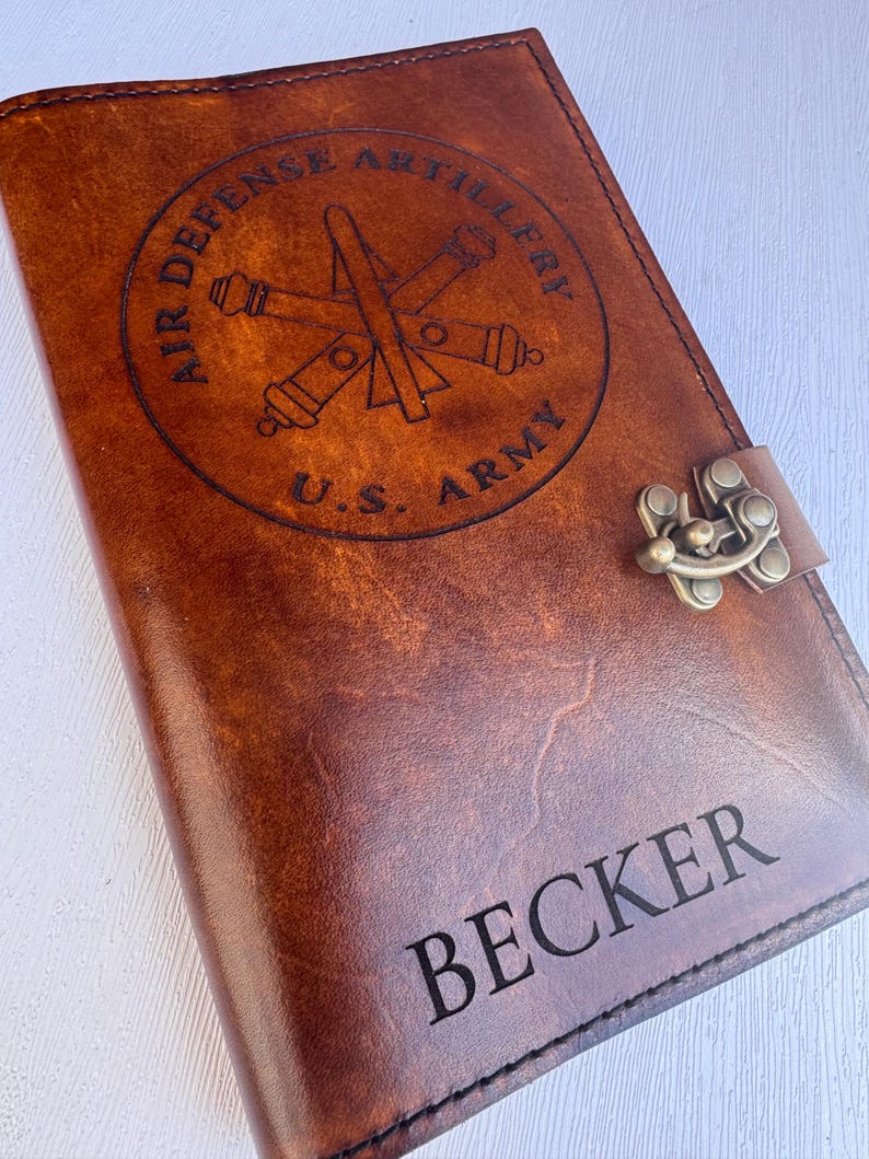 May include: A brown leather journal with the text "BECKER" engraved at the bottom. The cover features a circular emblem with the words "AIR DEFENSE ARTILLERY" and "U.S. ARMY" surrounding a crossed cannon and missile design. A metal clasp secures the journal.