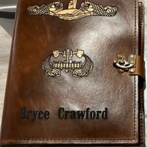 May include: A brown leather journal with a personalized inscription "Bryce Crawford". The journal features two gold embossed submarine insignia, one with a trident and the other with a submarine.