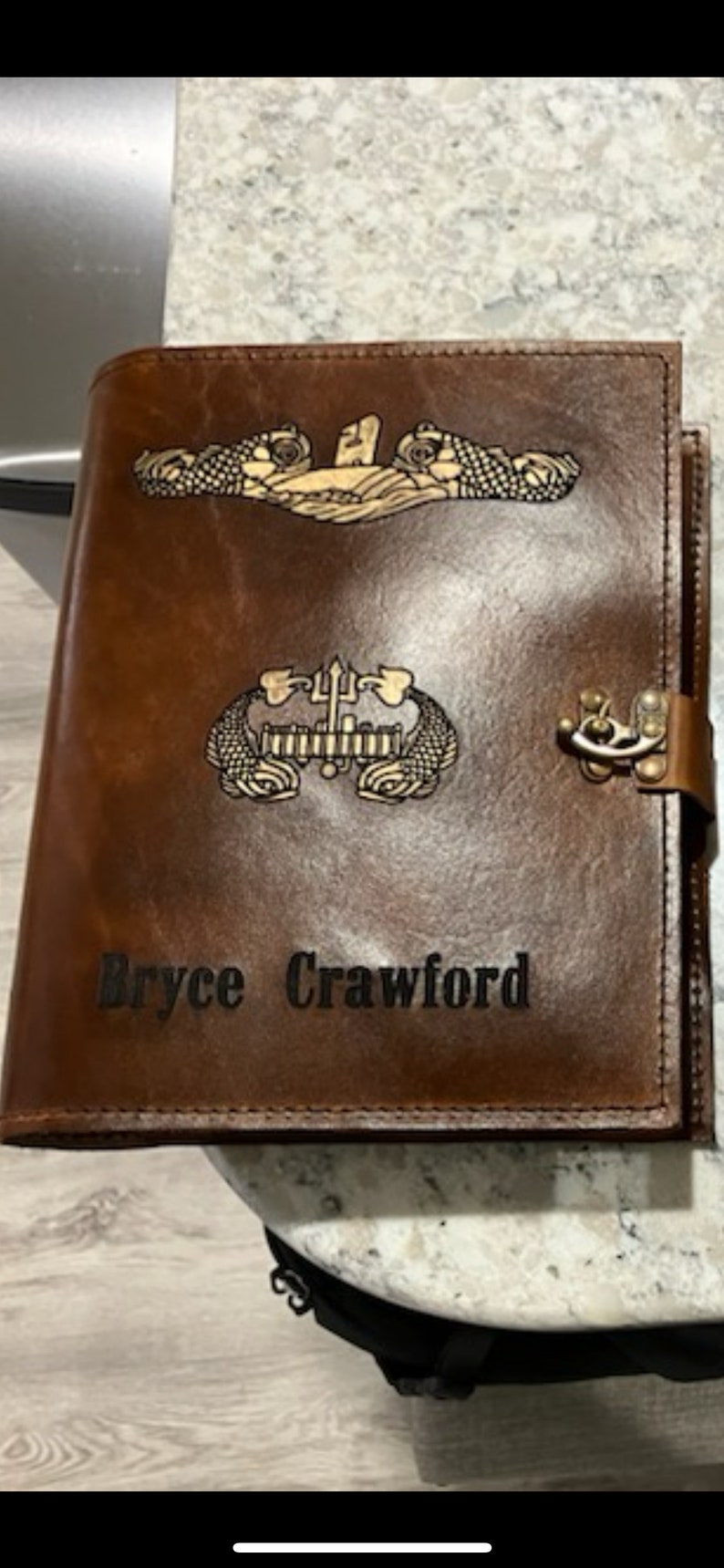 May include: A brown leather journal with a personalized inscription "Bryce Crawford". The journal features two gold embossed submarine insignia, one with a trident and the other with a submarine.