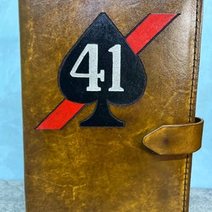 K&ouml;nnte beinhalten: Ein braunes Lederjournal mit einem schwarzen Pik mit der Nummer 41 in Wei&szlig; und einem roten Diagonalstreifen. Das Journal ist mit dem Wort "SHADY" auf der Vorderseite gepr&auml;gt.