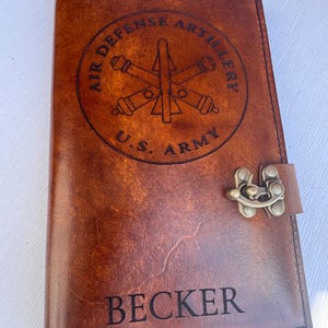 May include: A brown leather journal with a metal clasp. The cover features a circular emblem with the text "AIR DEFENSE ARTILLERY" and "U.S. ARMY". The name "BECKER" is engraved at the bottom of the cover.