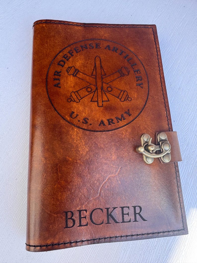 May include: A brown leather journal with a metal clasp. The cover features a circular emblem with the text "AIR DEFENSE ARTILLERY" and "U.S. ARMY". The name "BECKER" is engraved at the bottom of the cover.