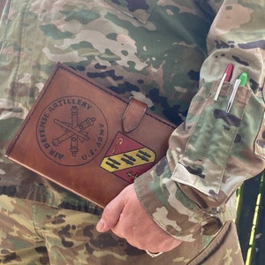May include: A brown leather journal with the words "AIR DEFENSE ARTILLERY" and a U.S. Army emblem. The journal is held by a person wearing a camouflage uniform. Two pens are in the uniform pocket.