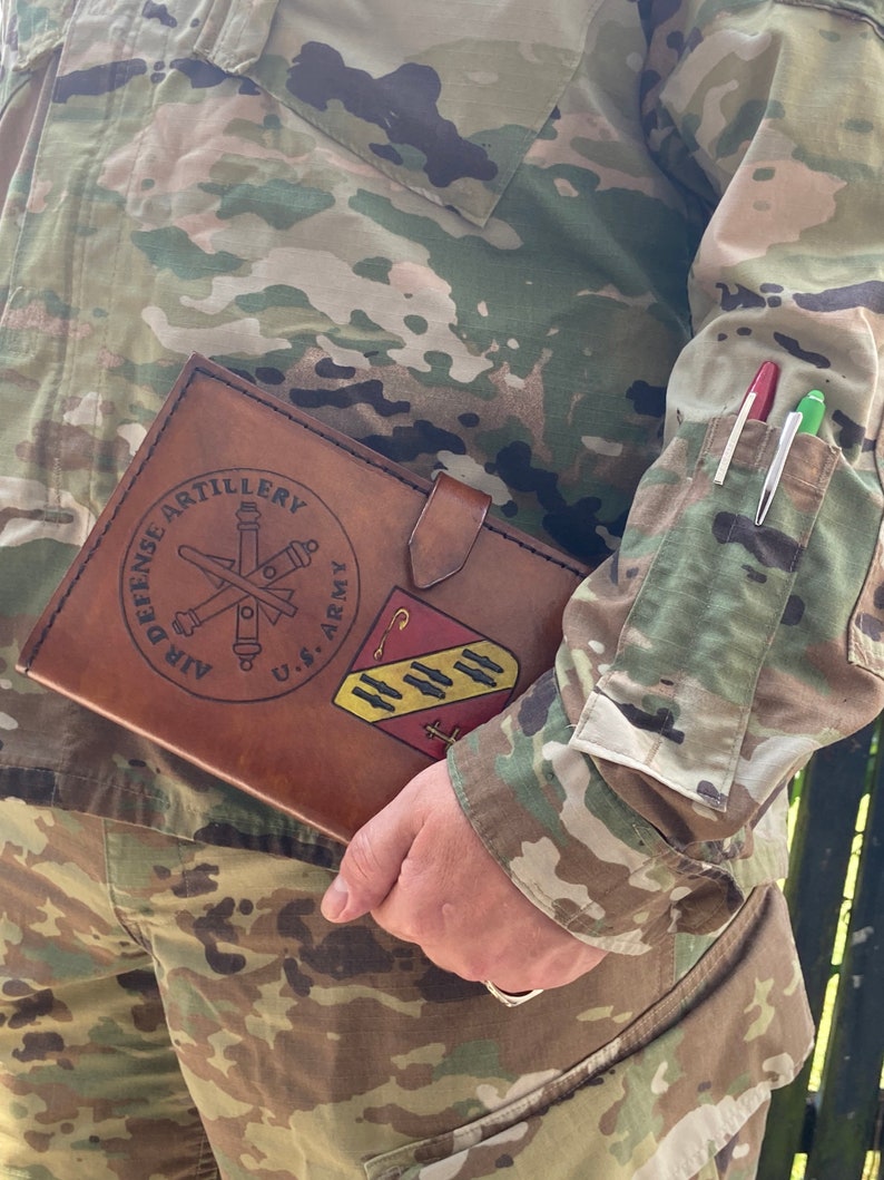 May include: A brown leather journal with the words "AIR DEFENSE ARTILLERY" and a U.S. Army emblem. The journal is held by a person wearing a camouflage uniform. Two pens are in the uniform pocket.