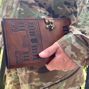 May include: A brown leather journal with a castle design and the word "MALTA" embossed on the cover. The journal has a metal clasp closure and is held against a camouflage background.