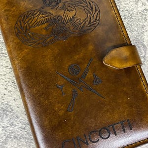 K&ouml;nnte beinhalten: Ein braunes Lederjournal mit einer personalisierten Inschrift "CINCOOTTI" und einem gravierten Bild eines milit&auml;rischen Abzeichens mit einem Vogel und einem Kranz.