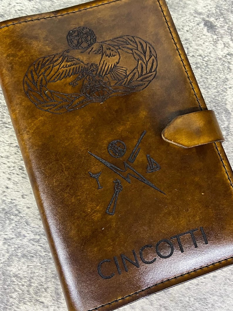 K&ouml;nnte beinhalten: Ein braunes Lederjournal mit einer personalisierten Inschrift "CINCOOTTI" und einem gravierten Bild eines milit&auml;rischen Abzeichens mit einem Vogel und einem Kranz.
