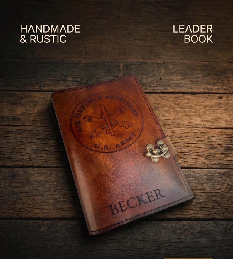 May include: A handmade, rustic leather journal with a brass clasp. The cover is embossed with an Air Defense Artillery emblem and the text "U.S. Army". The name "Becker" is also visible. The journal is set against a dark wood background.