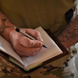 May include: A person with tattoos on both arms is writing in a brown leather bound journal with a silver pen.