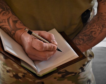 Puede incluir: Una persona con tatuajes en ambos brazos está escribiendo en un diario encuadernado en cuero marrón con una pluma plateada.