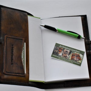 May include: A brown leather journal with a pen and business card inside. The journal has a pocket on the front cover and a strap to keep it closed. The business card says "Js Original Leather" and has a website address.