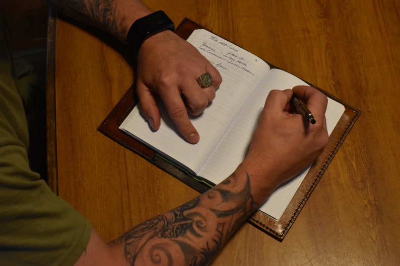 May include: A brown leather journal with lined pages is open on a wooden surface. A person is writing in the journal with a pen. The journal is held open by the person's hands. The person is wearing a ring and has tattoos on their arm.