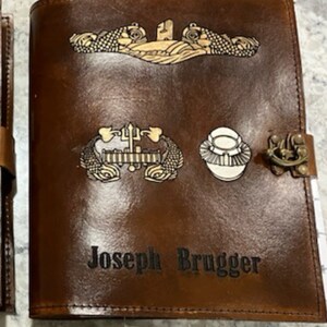 May include: A brown leather journal with a gold embossed design. The journal has a clasp and the name "Joseph Brugger" is embossed on the front.