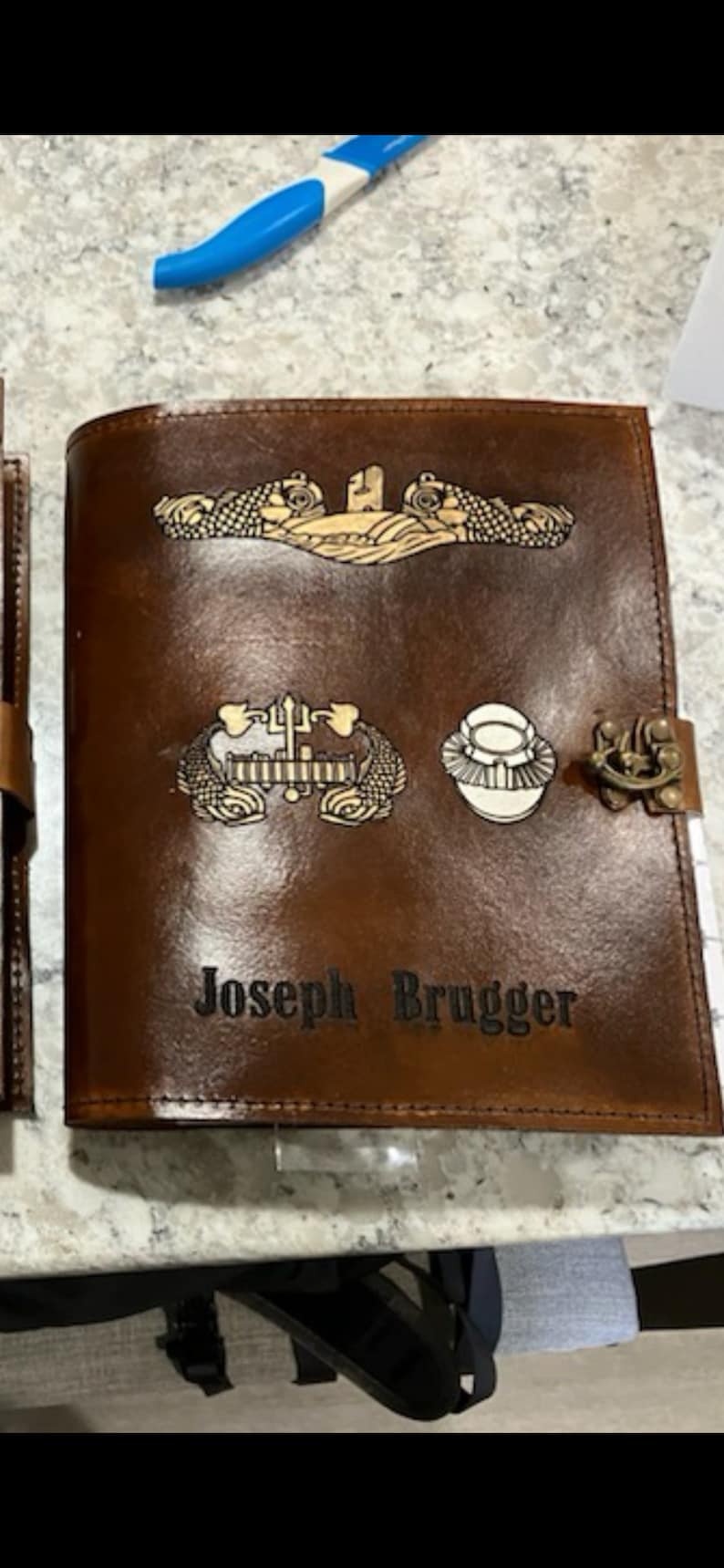 May include: A brown leather journal with a gold embossed design. The journal has a clasp and the name "Joseph Brugger" is embossed on the front.