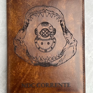 K&ouml;nnte beinhalten: Ein braunes Lederjournal mit einem Haimaul und einem Taucherhelm, die auf die Vorderseite graviert sind. Der Text "NDC CORRENTE" ist unter dem Bild graviert.