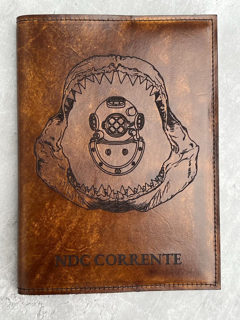 K&ouml;nnte beinhalten: Ein braunes Lederjournal mit einem Haimaul und einem Taucherhelm, die auf die Vorderseite graviert sind. Der Text "NDC CORRENTE" ist unter dem Bild graviert.