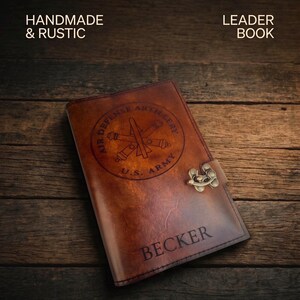 May include: A handmade, rustic leather journal with the text "Air Defense Artillery U.S. Army" and the name "Becker" embossed on the cover. The journal has a metal clasp and is set against a dark wood background.