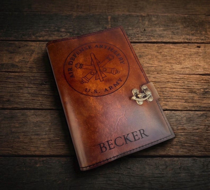 May include: A brown leather journal with a decorative clasp. The cover features a circular emblem with the text "AIR DEFENSE ARTILLERY U.S. ARMY" and the name "BECKER" engraved below the emblem.