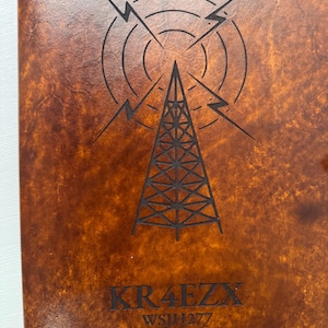 May include: A brown leather notebook cover with a detailed etched design. The design features a radio tower, concentric circles, and lightning bolts. The text "KR4EZX" and "WSJH277" are etched below the design.