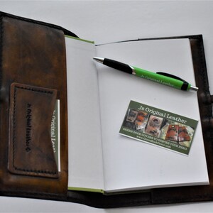May include: Open brown leather journal with a pen and business card. The journal has a pocket for cards and a strap closure. The pen is green and black. The business card reads "Js Original Leather".