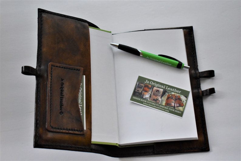 May include: Open brown leather journal with a pen and business card. The journal has a pocket for cards and a strap closure. The pen is green and black. The business card reads "Js Original Leather".