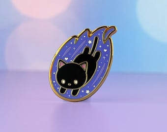 May include: A black cat pin with a gold edge, set against a purple and blue backdrop. The pin features a black cat inside a blue flame shape with stars and hearts. The pin has the text "Meow Amor Creative" and "Cynthia Tizcareno".