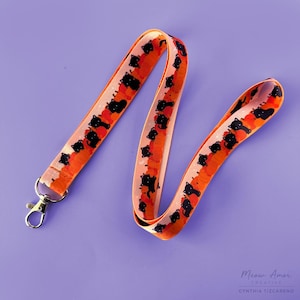 Könnte beinhalten: Ein Schlüsselband mit einem sich wiederholenden Muster aus schwarzen Katzen-Silhouetten auf orangefarbenem und pfirsichfarbenem Hintergrund. Das Schlüsselband hat einen silberfarbenen Metallverschluss und einen Karabinerhaken. Auf violettem Hintergrund.
