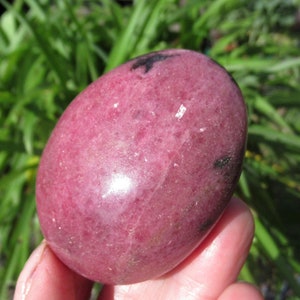 May include: A smooth, polished pink and black gemstone, shaped like an egg. The stone has a glossy finish and is held in a hand.