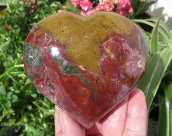 May include: A heart-shaped stone with a polished surface. The stone features a mix of colors, including deep red, brown, green, and yellow. The heart is held in a hand, with a background of pink flowers and green plants.