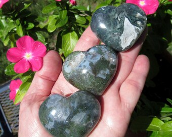 May include: Three green moss agate heart shaped stones in a hand. The stones are polished and have a natural green and white pattern.
