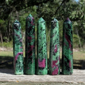 May include: Five green, black, and pink gemstone points, also known as zoisite, are arranged in a row on a wooden surface.
