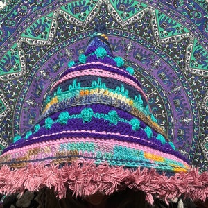 May include: A colourful crocheted hooded hat with a pink fringe. The hat is made with a variety of colours, including blue, purple, green, and pink. The hat is being worn by a person, but the person's face is not visible.