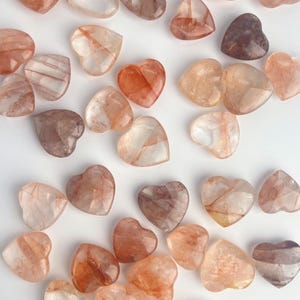 May include: A collection of heart-shaped gemstones in varying shades of red, brown, and clear. The stones are translucent, with visible internal patterns. The arrangement is scattered, showcasing the different colors and textures of the stones.