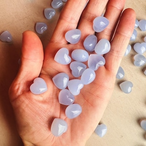 May include: A collection of small, heart-shaped, pale blue gemstones. The smooth, polished stones are held in an open hand, with several more scattered around. The gemstones have a translucent quality, catching the light.