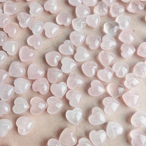Pode incluir: Uma coleção de pequenas pedras de quartzo rosa em forma de coração. As pedras variam em tons de rosa e estão espalhadas sobre uma superfície rosa claro. As pedras translúcidas têm uma aparência polida, refletindo a luz.