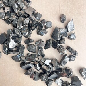 May include: A collection of irregularly shaped, shiny black obsidian stones scattered across a light tan surface. The stones vary in size and have a reflective, glassy appearance, with some showing subtle variations in texture and color.
