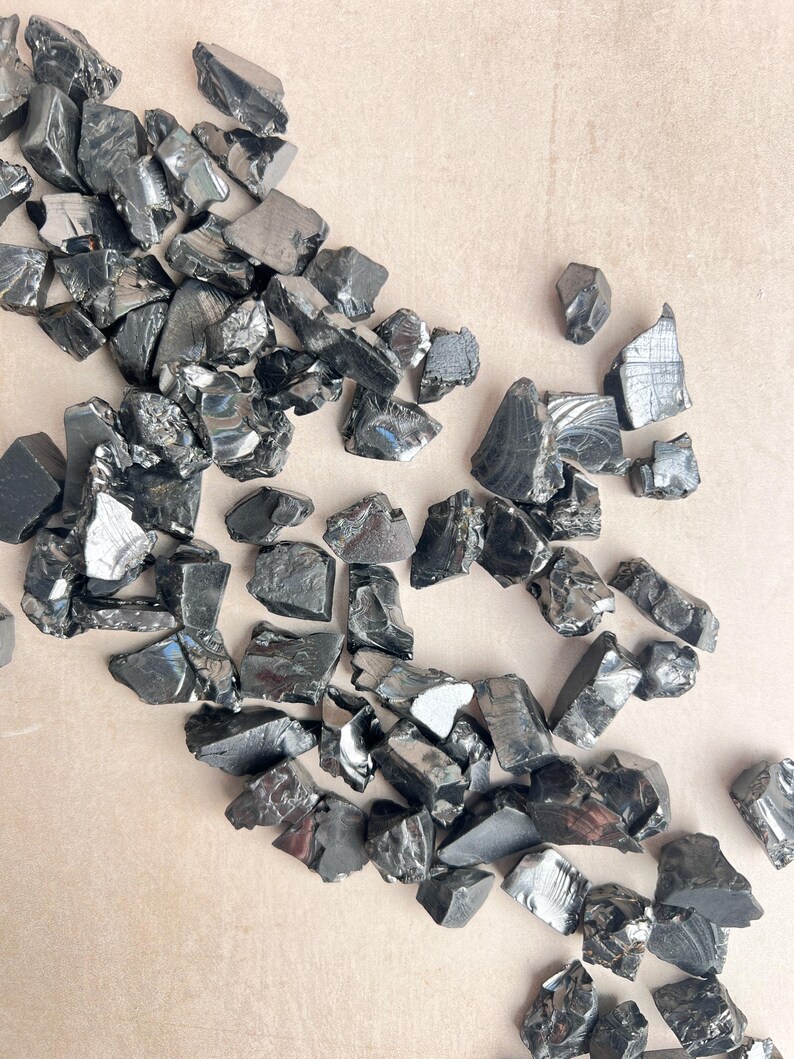May include: A collection of irregularly shaped, shiny black obsidian stones scattered across a light tan surface. The stones vary in size and have a reflective, glassy appearance, with some showing subtle variations in texture and color.