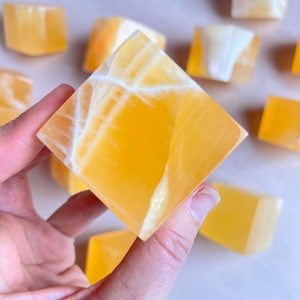 Puede incluir: Una mano sostiene un gran cristal de calcita dorada tallado en forma cuadrada con vetas blancas. Varios cubos de calcita más pequeños están dispersos en el fondo, todos con un color y patrón similares. Los cristales tienen una cualidad translúcida.