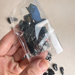 May include: A clear plastic bag filled with several pieces of black obsidian stones. The stones are irregularly shaped and have a glossy appearance. Some stones are outside the bag. The bag is held by a person.