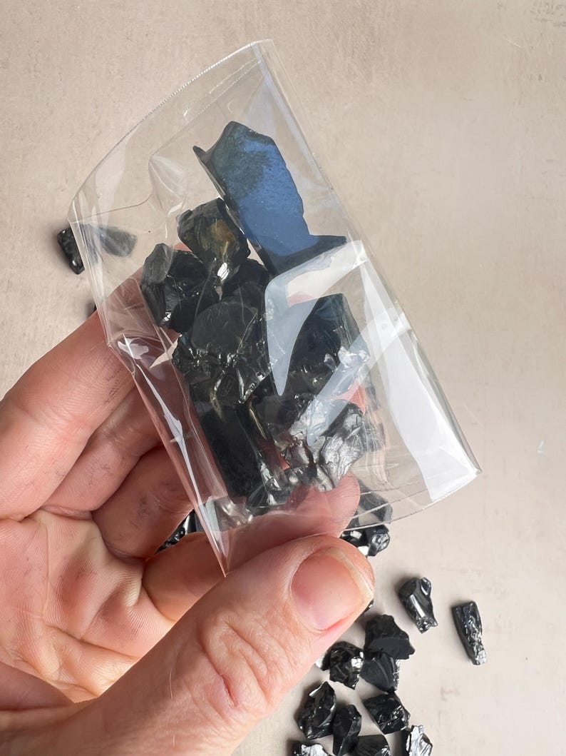 May include: A clear plastic bag filled with several pieces of black obsidian stones. The stones are irregularly shaped and have a glossy appearance. Some stones are outside the bag. The bag is held by a person.