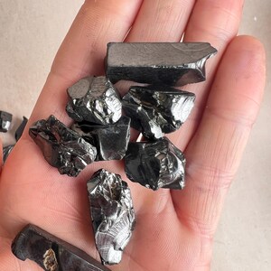 May include: A handful of shiny, black hematite stones. The stones have irregular shapes and a metallic sheen, reflecting light. The hematite pieces vary in size and texture, with some appearing smooth and others more jagged.