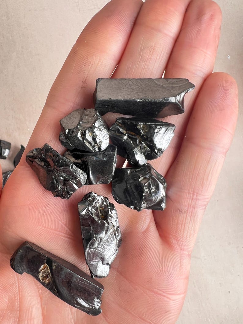 May include: A handful of shiny, black hematite stones. The stones have irregular shapes and a metallic sheen, reflecting light. The hematite pieces vary in size and texture, with some appearing smooth and others more jagged.