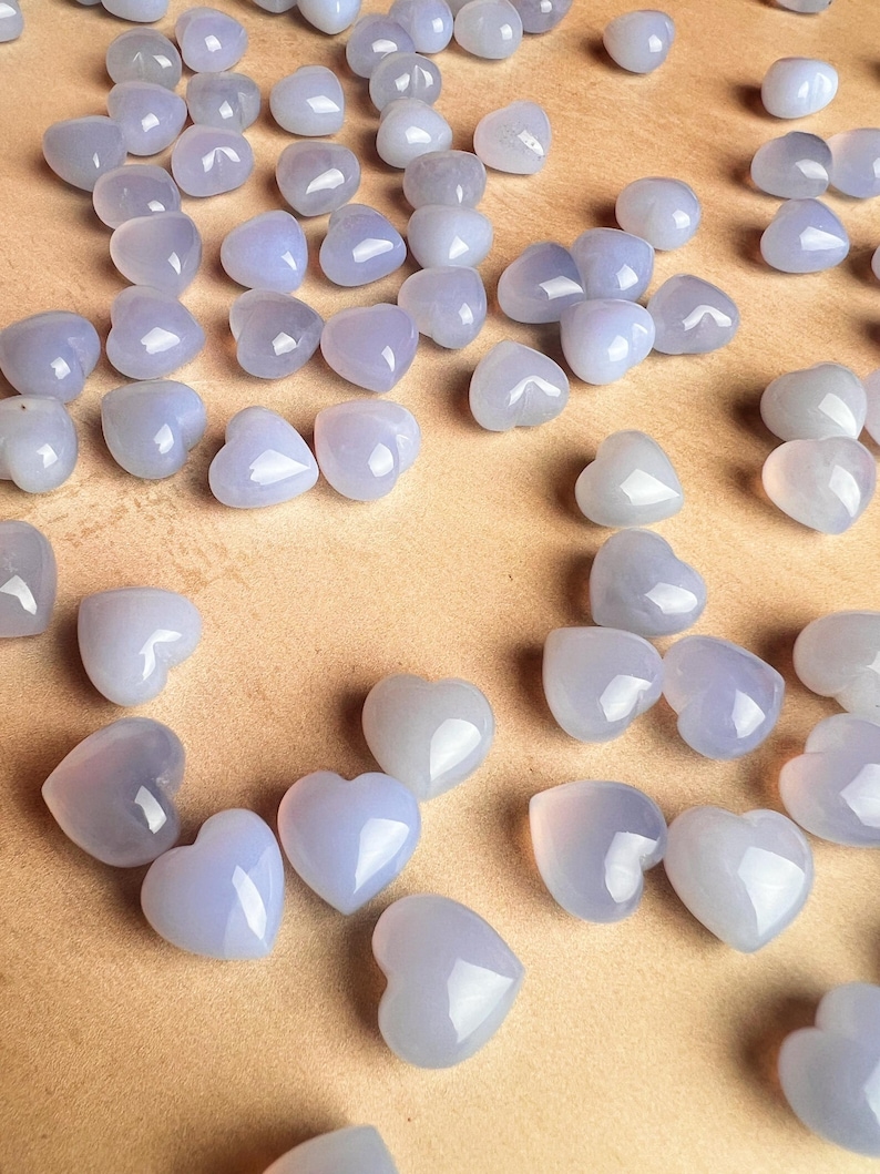 Puede incluir: Una colecci&oacute;n de peque&ntilde;as piedras en forma de coraz&oacute;n en tonos azul p&aacute;lido y lavanda. Las piedras pulidas est&aacute;n dispersas sobre una superficie marr&oacute;n claro, creando una est&eacute;tica suave y rom&aacute;ntica. Estas piedras en forma de coraz&oacute;n son ideales para la fabricaci&oacute;n de joyas o manualidades.